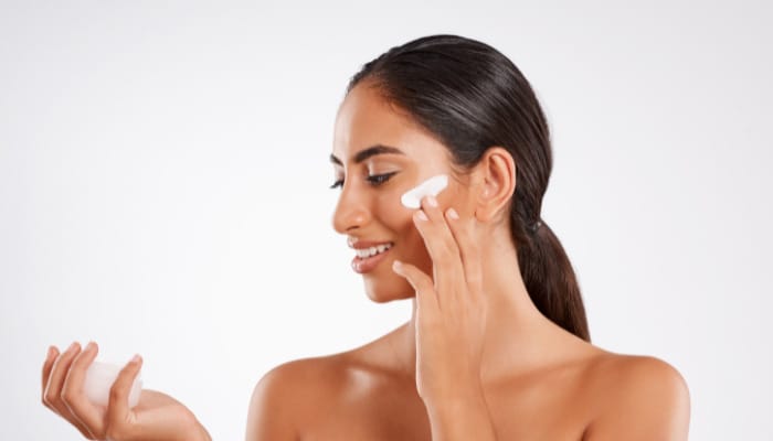 Model holding skincare jar, applying cream to cheek